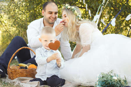 Little blond haired toddler boy, groom lying on knees of bride in wedding dress. Outdoor wedding scene in sunny summer dress. Young beautiful family has a picnic in summer natureの写真素材