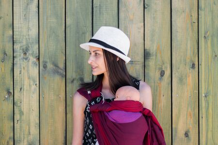 Almaty, Kazakhstan - July 08, 2019: Young beautiful babywearing mother carry her newborn baby in a ring sling on a rural green wooden wall background.のeditorial素材