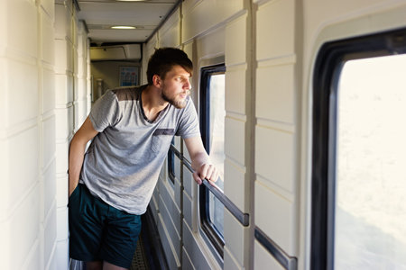 Handsome young caucasian man with a beard, looks outside of the train's window, looking at the...