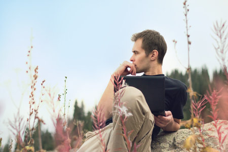 Handsome young concentrated man freelancer working on a laptop outdoors in mountains. Freelance...