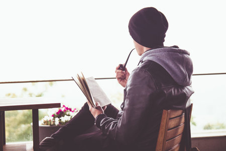 Back view on young black haired man witn piercing reeding a book and smoking tabacco pipe in outdoor...