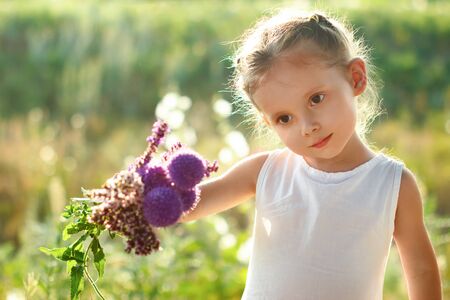 Little cute girl in white dress with a summer bouquet of wildflowers in hand walks in field. Selective soft focusの写真素材