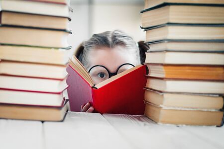 Serious little girl in round shaped glasses practicing reading between books pile. Selective soft focus. Primary education conceptの写真素材