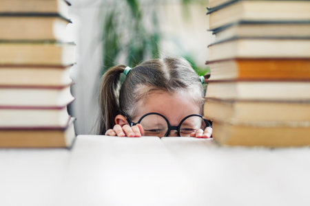 Little girl in big glasses with cute pony tails try to hide under a table from reading a pile of...