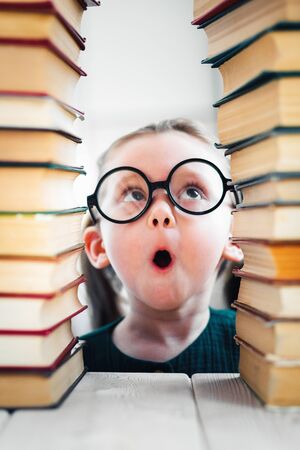 Shocked little blonde kid girl 4-6 years old in glasses in library among many books, children studio close-up portrait. Kid education concept.の写真素材