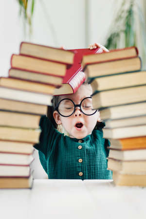 Cute little girl in round shape glasses and green dress behind arch made of books. Reading and kids education concept.の写真素材