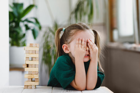 Cute caucasian little girl with ponytails closing her face with her hands, a wooden  tower standing...