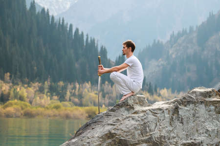 A wise man sits on a stone and holds a sword in his hands. Mountain Lake background.の写真素材