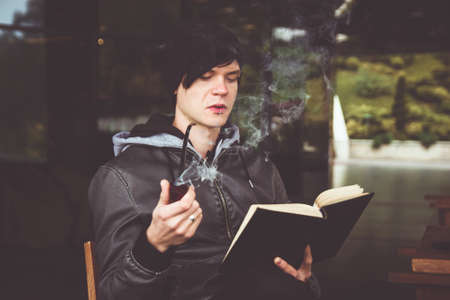 Young black haired man witn piercing reeding a book and smoking tabacco pipe in outdoor cafeの写真素材
