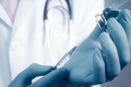 Close up view on syringe and glass ampoule in hands dressed in blue rubber gloves, white robe dressed doctor on blurred background.の写真素材