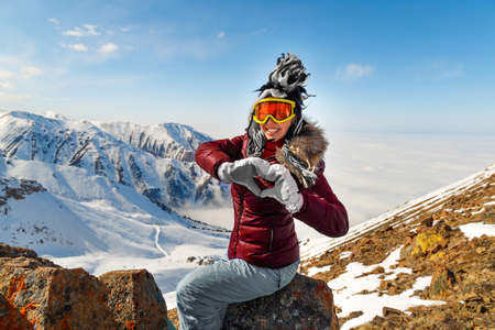 Funny young woman making a heart shape with her hands, sitting over snowy mountain peaks. Winter traveling scene, wanderlust concept.の写真素材