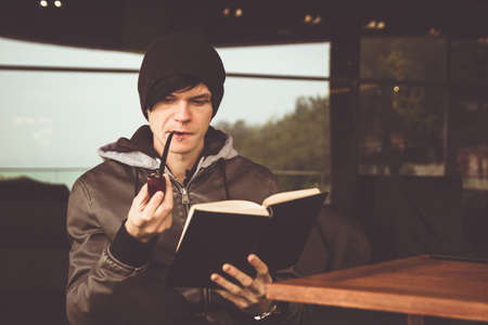 Back view on young black haired man witn piercing reeding a book and smoking tabacco pipe in outdoor cafe. Brown tinted photoの写真素材