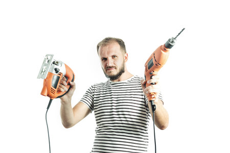 A serious man in a striped T-shirt, holds a drill and an electric jigsaw in his hands. Isolated on...