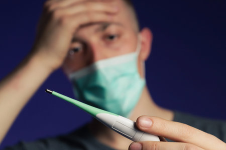 Caucasian sick man in a medical gauze mask with palm on his head holding thermometer in other hand, fever conceptの写真素材