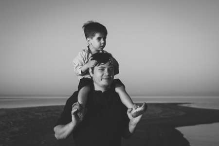 Black and white portrait of a cute boy 3 years old on his fathers neck walking on the beach in sunset lightingの写真素材