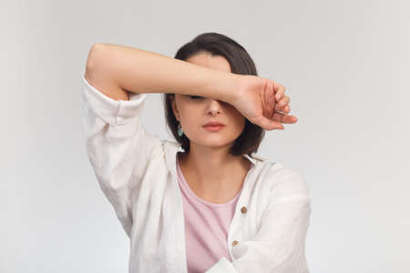 High-key studio portrait of a young woman in light clothes covering her eyes with her handの写真素材