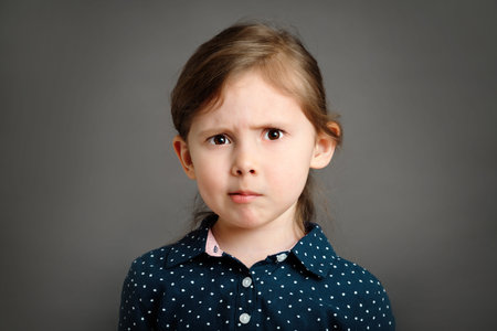Perplexed little caucasian kid girl 4 -6 years old in a blue dress with polka dots on gray...