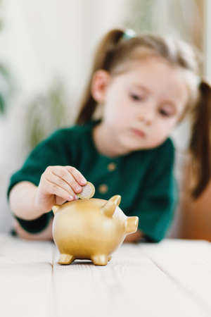 Close up view on child hand, she putting a coin in gold color piggy bank. Verticalの写真素材