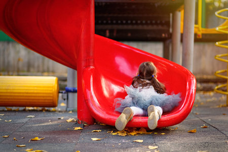 A cute girl in skirt tutu is riding a playground slide. Lifestyle outdoor portrait of a child.の写真素材