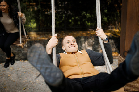 Young cheerful man swinging on a swing with legs raised high with his lover in the background in a forest park. High quality photoの写真素材