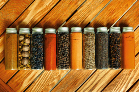 Different multi-colored indian spices in transparent jars lie on a wooden background.