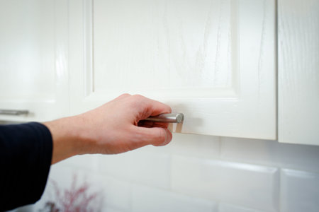 A man's hand is going to open the door of a hinged kitchen cabinet.