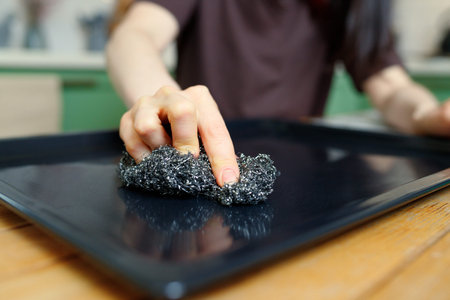 Close-up on a hand with a metal sponge that cleans a baking sheet from the oven.