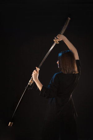 A young beautiful woman in a black kimono draws a Japanese katana from its scabbard, back view.