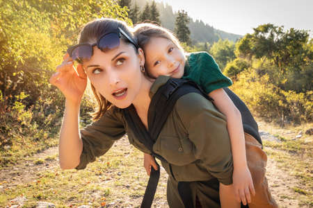 Happy preschool kid girl in babywearing sling at back her mother in hiking outdoor.の写真素材