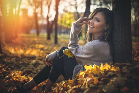Young happy mixed race woman sitting near tree in autumn park at sunset looking at camera.の写真素材