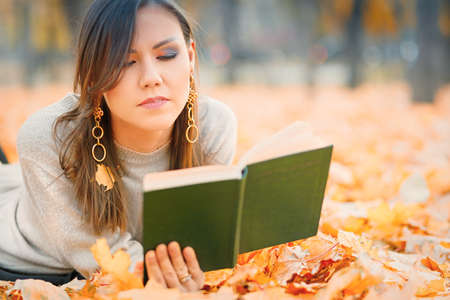 Central asian woman lying on fall foliage and reading book in park.の写真素材
