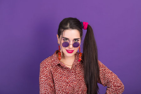 Smiling young brunette woman in 90s style with round shape sunglasses on purple background.