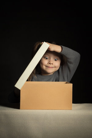 Emotional happy and surprised little girl opens a long-awaited package with a gift, copy space on cardboard box, vertical.の写真素材