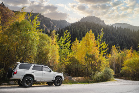 A white crossover 4x4 car driving on the countryside asphalt road against a autumn forest in mountains, sky with clouds and a beautiful sunset.の写真素材