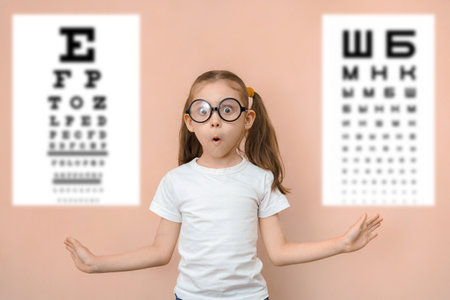 Surprised girl 7 years old in round glasses against the background of blurry tables for checking visual acuity.の写真素材
