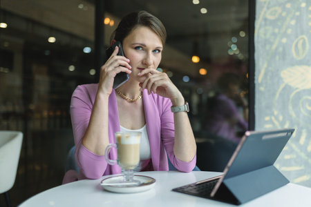 Young caucasian woman pensively looking at the camera while talking on a mobile phone while sitting in a cafe with a cup of latte.の写真素材
