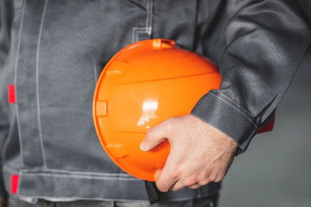 Orange safety helmet is in the hands of a construction worker, close up, construction site safety concept.の写真素材