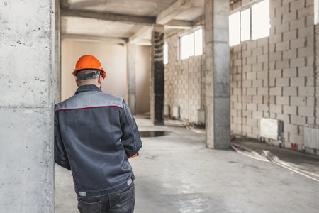 A construction worker in a helmet stands with his back to the camera, leaning on a reinforced...