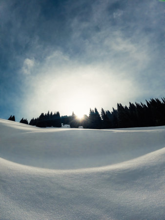 A wintry scene with snowcovered mountain slopes, trees in the background, and the sun peeking through the clouds, creating a picturesque natural landscape.の写真素材