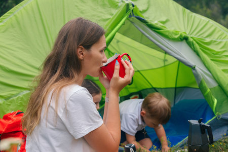 A woman willingly enjoys a refreshing drink while her children happily play in a vibrant tent during a camping tripの写真素材