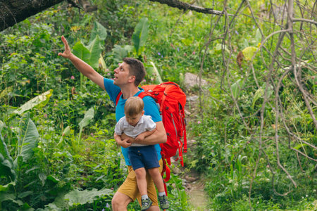 A father joyfully carries his child on his back, gazing at the vibrant forest as they start their hiking adventureの写真素材