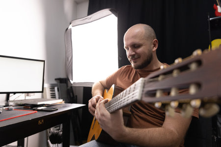 A man focuses on playing an acoustic guitar in a cozy home music studio setup with soft lighting.