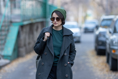 Portrait of a young woman in a green beanie, round sunglasses, and a grey coat walking down a city...