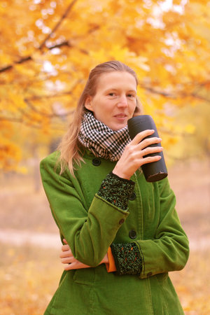 Girl in green coat with a mug in her hand standing near autumn treeの写真素材