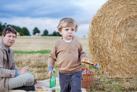 Father and beautiful blond toddler boy making evening picnic on golden straw fieldの写真素材