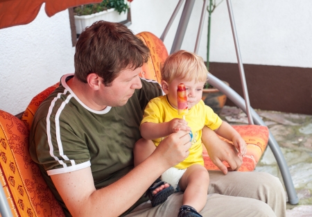 Young man and little baby boy eating colorful ice in summer, outdoorsの写真素材