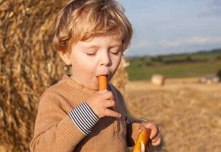 Lovely little boy of two years with pretzel and sausage on hay field, Germanyの写真素材