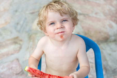 Adorable little toddler boy with blond hairs eating watermelon in summergardenの写真素材