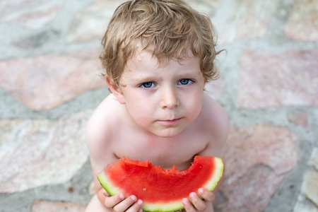 Adorable little toddler boy with blond hairs eating watermelon in summergardenの写真素材