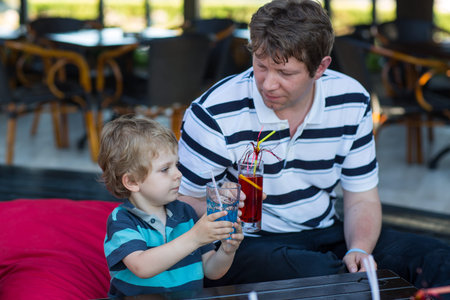 Father and little son sitting in cafe with cocktail on sunny summer day and drinking